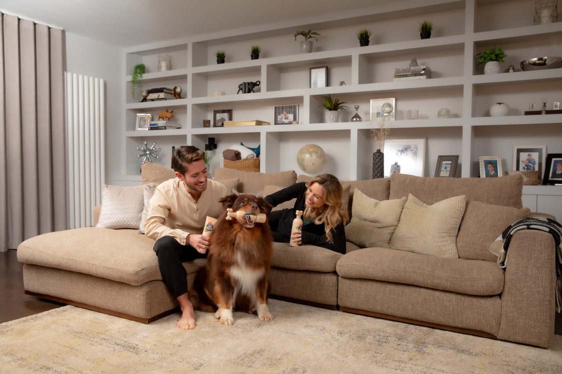 Young adult couple smiling and petting their collie who has Sensitive soul shampoo bottle in his mouth
